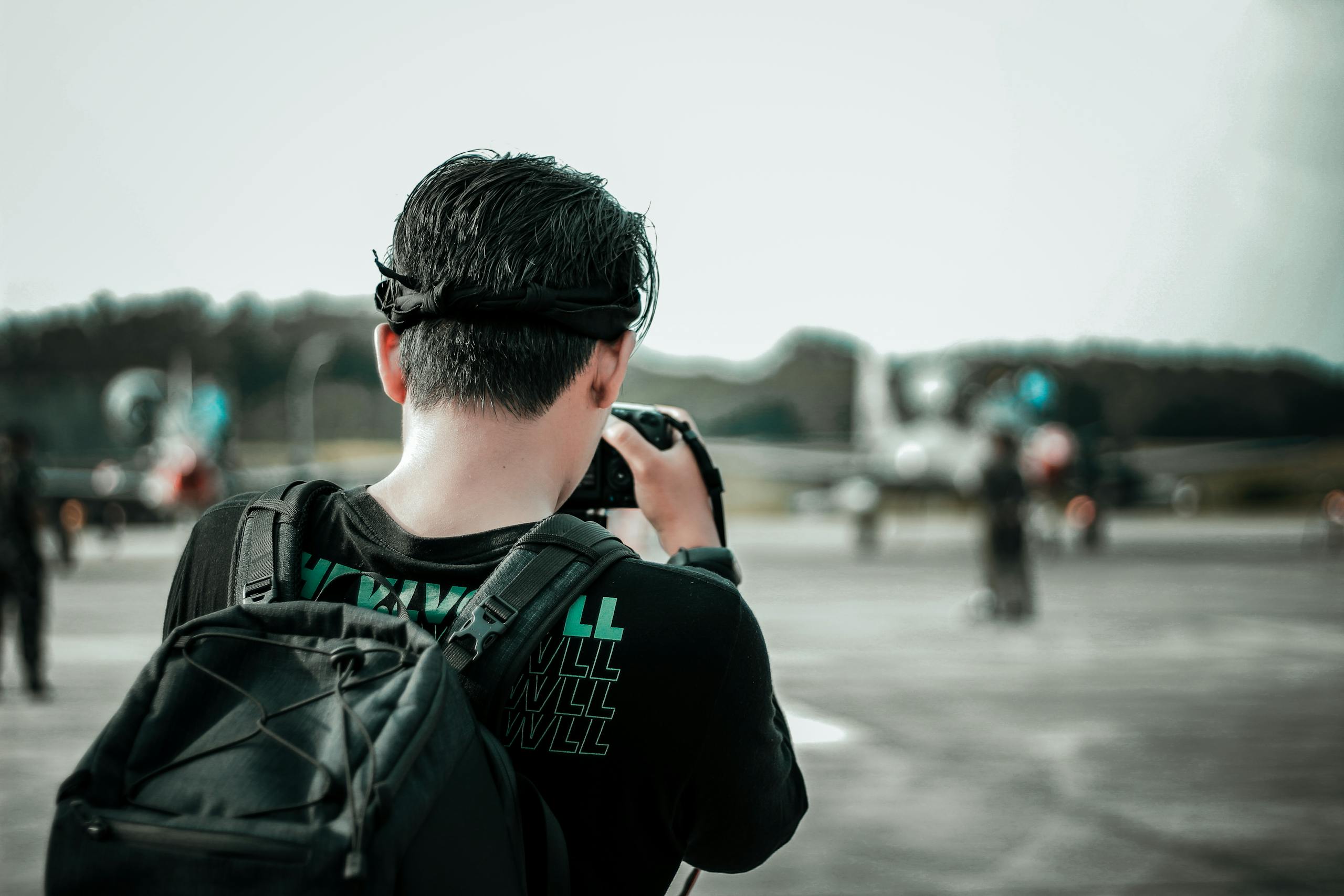 A man with a backpack photographs airplanes on an outdoor runway during the day.